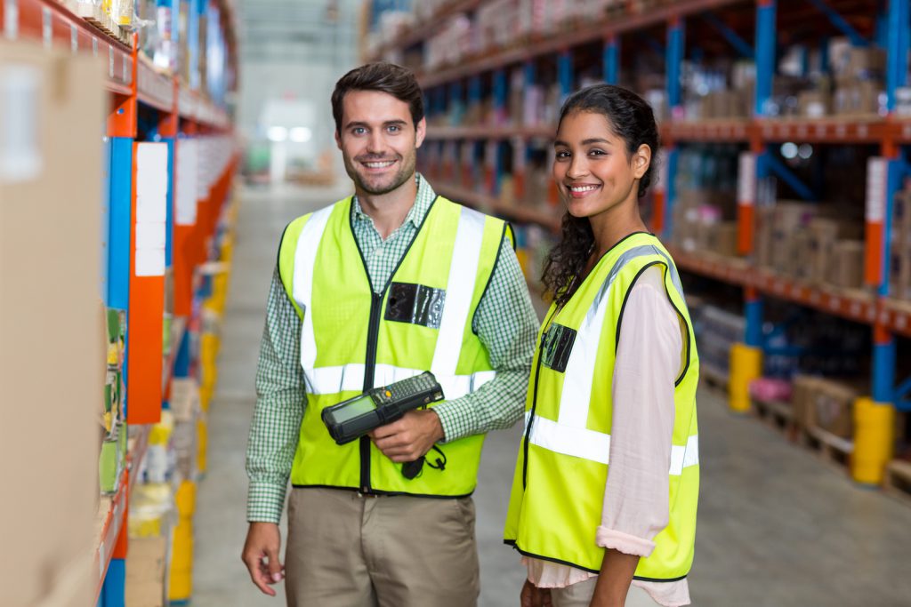 Two warehouse employees smiling and wearing bright yellow safety vests.