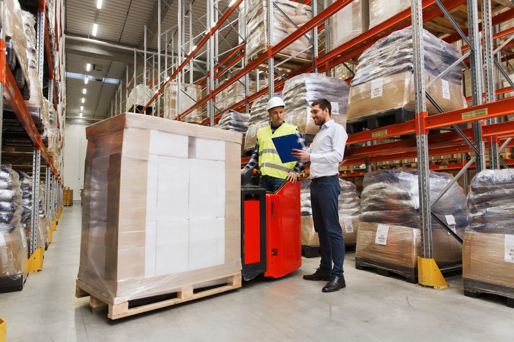 Two workers discussing information on a clipboard in a storage warehouse near a pallet.